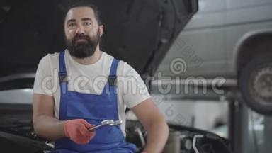 在一家汽车修理店里，有着迷人的胡须的机械师摆姿势，扳手坐在一辆破车的引擎盖上。